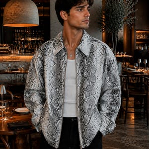 Puede incluir: Una chaqueta de manga larga con estampado de piel de serpiente en gris y blanco, cuello en punta y cremallera frontal completa. La chaqueta se lleva sobre una camiseta de tirantes blanca lisa, mostrando un corte estructurado y clásico.