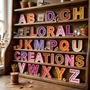 Könnte beinhalten: Ein Holzregal zeigt eine Sammlung dreidimensionaler Buchstaben von A bis Z, die jeweils mit komplizierten Blumenmustern und leuchtenden Farben versehen sind. Die Buchstaben sind in Reihen angeordnet, durchsetzt mit kleinen Topfpflanzen und Lavendel.
