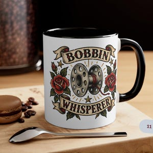 May include: A white ceramic mug with a black handle and rim, featuring a graphic of a sewing bobbin surrounded by red roses and green leaves. The text "BOBBIN WHISPERER" is printed in a banner style above and below the bobbin. The mug is labelled 11 oz.