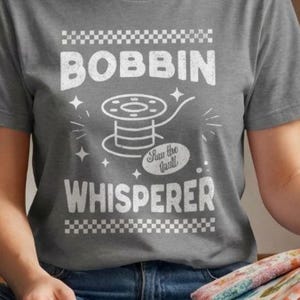 May include: A heather grey short-sleeve t-shirt featuring a white graphic print. The design includes a checkered border, the text BOBBIN WHISPERER, a sewing bobbin illustration, and the script text Sew the Vault surrounded by small stars.
