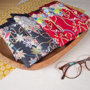 May include: A collection of rectangular fabric pouches featuring Japanese-inspired floral patterns in black and red, arranged in a wooden bowl alongside a pair of brown-rimmed spectacles. The pouches have gathered black fabric tops.