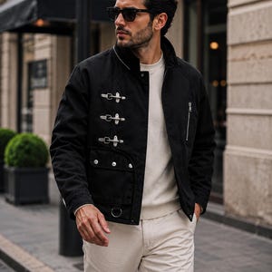 May include: A black jacket featuring three silver-toned metal toggle closures on the right side, a vertical zippered pocket on the left chest, a flap pocket with silver buttons, and a small metal ring detail at the bottom hem, worn over a cream sweater.