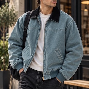 May include: A blue denim jacket with a black corduroy collar, a front zipper closure, and ribbed cuffs. The jacket is worn over a white crew neck shirt and paired with dark gray jeans. A black backpack strap is visible over the shoulder.