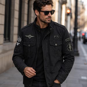 May include: A black military-style field jacket featuring a high collar, front button-flap pockets, and shoulder epaulets. The jacket is adorned with various embroidered patches, including a skull design on the sleeve and a winged emblem on the chest.