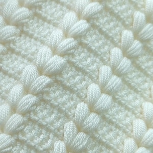 May include: A cream-colored crocheted fabric featuring a repeating pattern of diagonal rows of raised puff stitches alternating with sections of dense, flat single crochet stitches, creating a textured, uniform, and soft-looking textile surface.