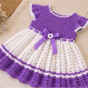May include: A crochet dress featuring a solid purple bodice with short ruffled sleeves and a white skirt with a diamond pattern. The waist is accented with a purple ribbon threaded through the fabric, tied in a bow with a small white flower.