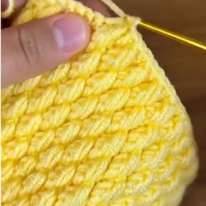 May include: A bright yellow crochet project featuring a dense, textured stitch pattern created with a metallic gold crochet hook. The yarn is worked in repeating diagonal rows, forming a consistent, raised surface across the entire piece.