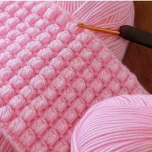 May include: A light pink yarn project featuring a textured bobble stitch pattern, worked with a crochet hook that has a black handle and a gold-colored metal shaft. The yarn is soft and pastel-toned, showing a consistent grid of raised stitches.