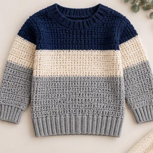 May include: A long-sleeved jumper featuring a colour-blocked design with a dark navy blue upper section, a cream-coloured middle band, and a light grey lower section. The garment has a textured waffle-knit pattern and ribbed cuffs and hem.