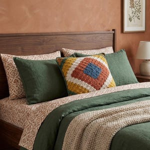 Könnte beinhalten: Ein Bett mit einem dunkelgrünen Leinen-Bettbezug und passenden Kissenbezügen, kombiniert mit einem cremefarbenen Laken mit braunem Blumenmuster. Ein quadratisches Häkelkissen mit Rautenmuster in Gelb, Weiß und Rostorange.