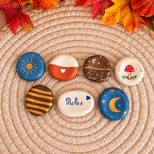 Peut inclure: Six galets ronds et lisses peints à la main, disposés sur deux rangées. Les motifs incluent un soleil bleu, un cœur bicolore orange, un imprimé léopard marron, un champignon rouge, des rayures dorées, le mot Relax et un croissant de lune bleu.