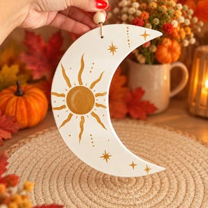Peut inclure: Un ornement en forme de croissant de lune blanc orné d'un soleil doré peint en son centre avec des rayons ondulés et de petites étoiles. L'ornement est suspendu par une ficelle avec une perle en bois au sommet, conçu pour être accroché.