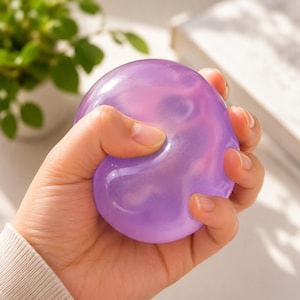 May include: A round, translucent purple stress ball with a smooth, glossy finish. The object is pliable and squishy, showing a soft, jelly-like texture that conforms to the shape of the grip. The material has a subtle, pearlescent sheen throughout.