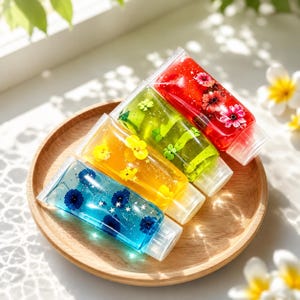 May include: Four clear squeeze tubes filled with colorful gel, each containing embedded dried flowers and small star-shaped glitter. The tubes are arranged diagonally, featuring blue, yellow, green, and red gel, each with a matching floral accent.