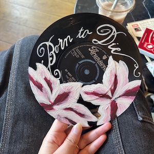 May include: A black vinyl record featuring hand-painted white and deep red flowers at the bottom. The text Born to Die is written in white cursive script across the top. The center label displays the Brunswick logo and song information.