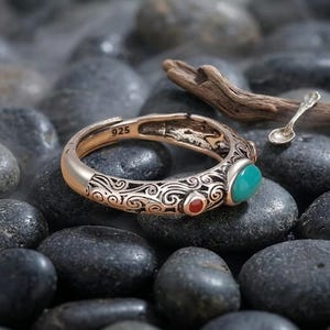 Peut inclure: Une bague argent&eacute;e &agrave; anneau ouvert orn&eacute;e de gravures complexes en volutes, d'une petite pierre ronde rouge et d'une pierre ovale plus grande de couleur turquoise. L'int&eacute;rieur de l'anneau porte l'inscription 925, indiquant de l'argent sterling.