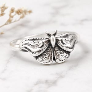 May include: A silver-toned ring featuring a detailed moth design on the front. The moth has textured wings with intricate engraved patterns and a smooth, rounded body. The band of the ring is plain and polished, connecting to the moth centerpiece.