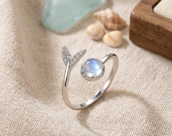 Peut inclure: Une bague ouverte argentée dotée d'une pierre cabochon ronde bleu irisé à une extrémité et d'un ornement en forme de queue de poisson incrusté de petites pierres claires scintillantes à l'autre. L'anneau est lisse et poli, offrant un design délicat.