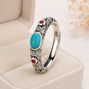Peut inclure: Une bague en argent orn&eacute;e d'une pierre turquoise ovale au centre, entour&eacute;e de deux petites pierres rouges rondes. L'anneau pr&eacute;sente des motifs m&eacute;talliques complexes en forme de spirales et porte l'inscription 925 sur sa face interne.
