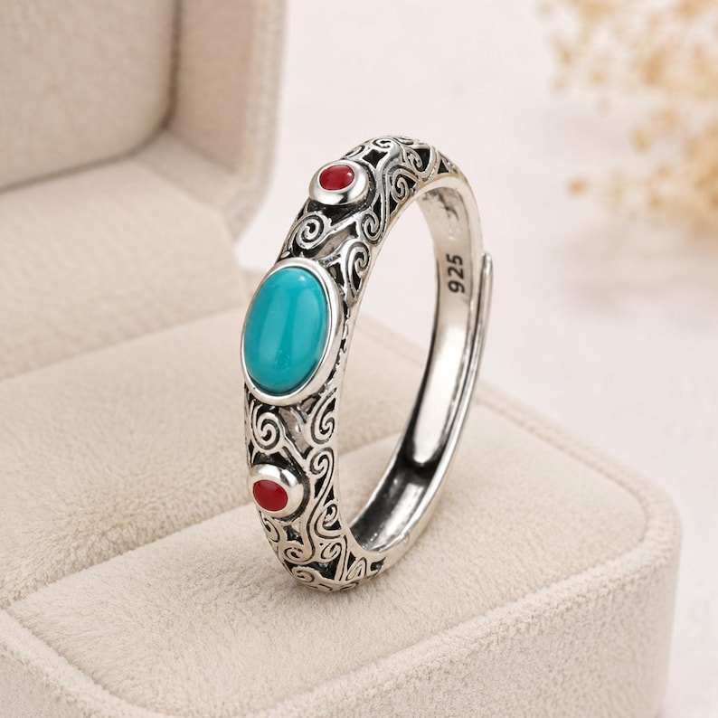 Peut inclure: Une bague en argent orn&eacute;e d'une pierre turquoise ovale au centre, entour&eacute;e de deux petites pierres rouges rondes. L'anneau pr&eacute;sente des motifs m&eacute;talliques complexes en forme de spirales et porte l'inscription 925 sur sa face interne.