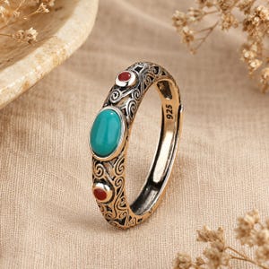 Peut inclure: Une bague en argent sterling avec un poin&ccedil;on 925, orn&eacute;e d'une pierre centrale ovale de couleur turquoise et de deux petites pierres rondes rouges. L'anneau est d&eacute;cor&eacute; de motifs de volutes complexes sur toute la surface m&eacute;tallique.
