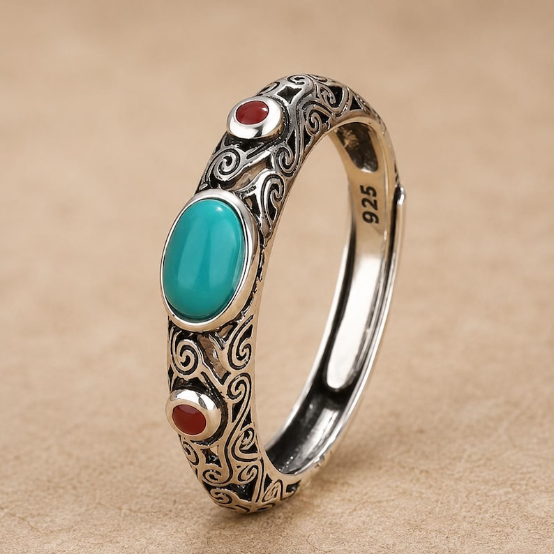 Peut inclure: Une bague en argent sterling avec un poin&ccedil;on 925, orn&eacute;e d'une pierre ovale de couleur turquoise au centre, entour&eacute;e de deux petites pierres rondes rouges. L'anneau pr&eacute;sente des motifs complexes de volutes en argent sur toute sa surface.