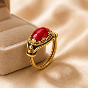 May include: A gold-toned ring featuring a smooth, oval-shaped red cabochon stone set in a decorative bezel with rope-like detailing and small scalloped edges. The band has a split design with a small spherical accent on each side of the setting.