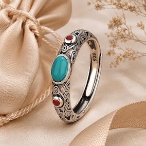 Può includere: Un anello in argento sterling con una pietra turchese ovale al centro, affiancata da due pietre rosse rotonde più piccole. La fascia è decorata con intricati motivi a volute ed è stampata con il numero 925 sulla superficie interna.