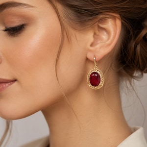 Pode incluir: Um brinco pendente de ouro com uma grande pedra preciosa facetada de cor vermelho-profundo e formato oval no centro. A pedra é rodeada por uma moldura de filigrana de ouro vazada com uma borda texturizada de contas, suspensa por um gancho de ouro.