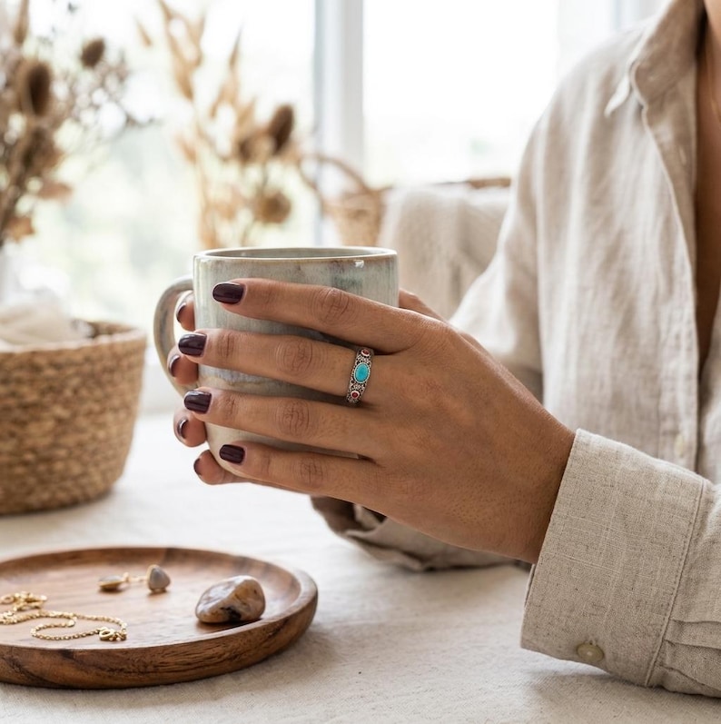 Peut inclure: Une bague en argent orn&eacute;e d'une pierre turquoise ovale centrale flanqu&eacute;e de petites pierres rouges, port&eacute;e au doigt. La bague pr&eacute;sente un anneau en m&eacute;tal travaill&eacute; avec des d&eacute;tails d&eacute;coratifs. L'objet est pr&eacute;sent&eacute; avec une tasse en c&eacute;ramique.