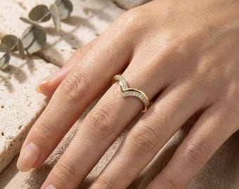 Peut inclure: Une bague dorée en forme de chevron ornée d'une rangée de petites pierres claires serties pavé sur la surface supérieure. L'anneau présente une finition polie et un design en V distinct qui repose sur le doigt, révélant éclat et brillance.