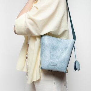 May include: A light blue bucket bag with a distressed texture and a long shoulder strap. The bag features a rectangular brand label on the front and a small, dangling shell-shaped charm attached to the side. The surface has a mottled finish.