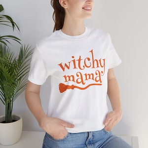 May include: A white short-sleeved t-shirt featuring the orange text "witchy mama" printed on the front. The text is accompanied by a graphic of a broomstick underneath the word "mama". The shirt has a crew neckline and a standard fit.