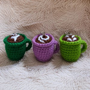 Könnte beinhalten: Drei gehäkelte Tassen-Schlüsselanhänger mit dunkelbraunen Garnoberteilen und weißen gestickten Latte-Art-Mustern. Die Tassen sind aus dunkelgrünem, hellviolettem und hellgrünem Garn gefertigt und jeweils mit einem silbernen Metallschlüsselring versehen.