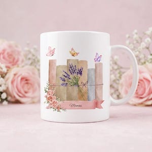 Könnte beinhalten: Ein weißer Keramikbecher mit einer Aquarellillustration von einem Stapel Bücher, die mit einer Schnur zusammengebunden sind, einem Strauß violetter Lavendel, rosa Rosen und drei kleinen Schmetterlingen. Ein rosa Banner trägt den Text Mamas.