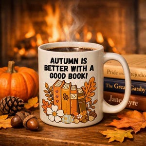Könnte beinhalten: Ein weißer Keramikbecher mit dem schwarzen Text "AUTUMN IS BETTER WITH A GOOD BOOK!" über einer farbenfrohen Illustration von gestapelten Büchern, Herbstblättern, einem kleinen Kürbis und einer Eichel. Der Becher hat einen Henkel und enthält ein dunkles Getränk.
