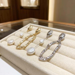 Puede incluir: Dos pares de pendientes colgantes con marcos de metal texturizado irregular y perlas barrocas blancas. Un par tiene un acabado en tono dorado pulido, mientras que el otro par tiene un acabado en tono plateado pulido, ambos con formas orgánicas.