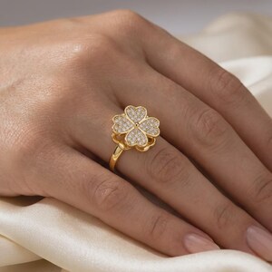 Op de afbeelding: Een goudkleurige ring met een klavertjevier-motief als middelpunt. Het klaverblad is ingelegd met talloze kleine, heldere, fonkelende steentjes die het gehele oppervlak van de vier blaadjes bedekken, wat zorgt voor een heldere uitstraling.