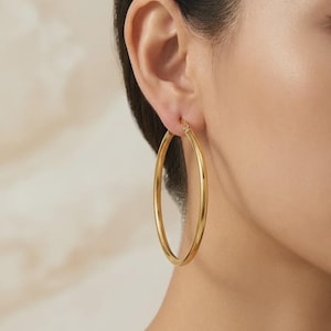 Pode incluir: Um brinco de argola grande em ouro polido com um perfil circular liso. O brinco apresenta um mecanismo de fecho de dobradiça na parte superior, prendendo o aro metálico contínuo. O design é simples e uniforme, com um acabamento metálico brilhante.