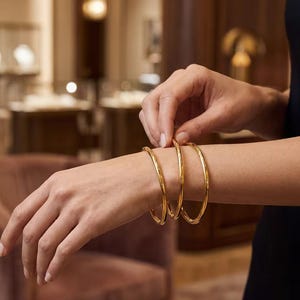 Puede incluir: Tres brazaletes rígidos de tono dorado pulido con un perfil liso y redondeado. Los brazaletes se llevan juntos en la muñeca, presentando un diseño circular continuo con un acabado metálico reflectante que capta la luz.