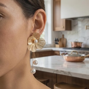 Puede incluir: Un pendiente de metal dorado con forma de flor y una perla blanca central. Dos hilos dorados finos cuelgan de la flor, cada uno terminado en una perla blanca de diferentes tamaños, creando un diseño colgante que descansa junto al rostro.