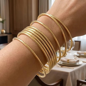 Puede incluir: Un conjunto de múltiples brazaletes finos de metal en tono dorado pulido usados juntos. Cada brazalete presenta una superficie lisa y redondeada con un diseño circular continuo, creando una apariencia uniforme y superpuesta en la muñeca.