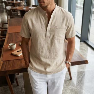 May include: A short-sleeved beige shirt with a mandarin collar, a partial button-down placket, and a single chest pocket. The shirt is paired with white trousers. The fabric has a visible textured weave, and the design features a straight hem.