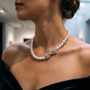 Puede incluir: Un collar que presenta una hilera de perlas blancas con un cierre plateado prominente en forma de dos motivos florales entrelazados incrustados con pequeñas piedras transparentes, usado sobre una prenda oscura de hombros descubiertos.