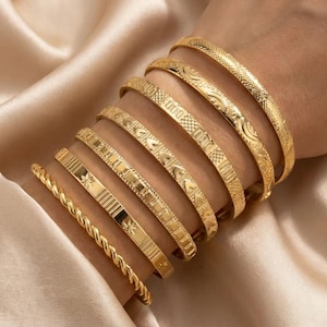 Puede incluir: Un conjunto de siete brazaletes de metal dorado con diversas texturas y patrones, incluyendo un diseño de cuerda trenzada, motivos de estrellas, cuadrados geométricos, formas de corazón y detalles de cuadrícula, usados juntos en la muñeca.