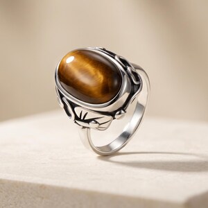 Puede incluir: Un anillo de plata con una gran piedra preciosa de ojo de tigre pulida de forma ovalada, engastada en una montura de metal decorativa. La banda es lisa y pulida, mientras que el engaste muestra intrincados detalles tallados.
