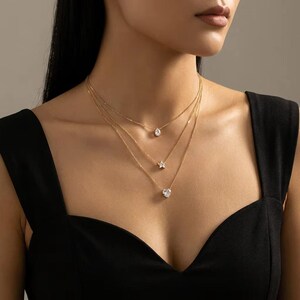 Puede incluir: Un collar de capas en tono dorado con tres cadenas finas de diferentes longitudes. Cada cadena sostiene un colgante de piedra transparente facetada en forma de gota, una estrella pequeña y un corazón, respectivamente, sobre la clavícula.