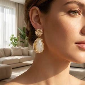 Puede incluir: Un pendiente dorado que presenta un perno circular con textura martillada conectado a un gran colgante de piedra irregular y facetada en tonos blanco y ámbar, enmarcado en un fino borde de metal dorado que cuelga del lóbulo.