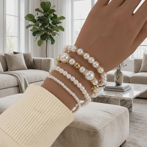 Puede incluir: Un conjunto de cuatro pulseras superpuestas con perlas artificiales blancas esféricas de varios tamaños, intercaladas con pequeñas cuentas metálicas doradas. Las pulseras se llevan en la muñeca y están hechas con un cordón elástico flexible.