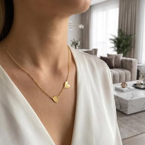 Puede incluir: Un collar de tono dorado con una cadena fina y dos pequeños dijes asimétricos en forma de corazón. Los dijes están colocados a lo largo de la cadena, con un corazón ligeramente más alto que el otro, creando un aspecto superpuesto.